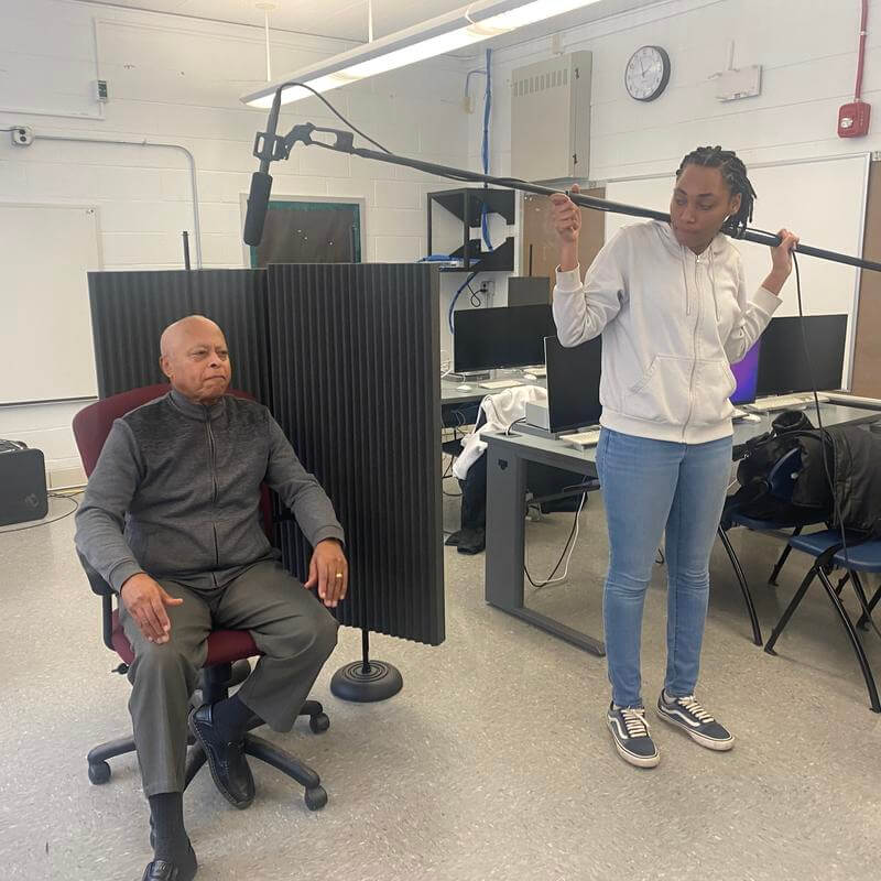 Audio Production student with sweatshirt holds mic in front of seated male subject
